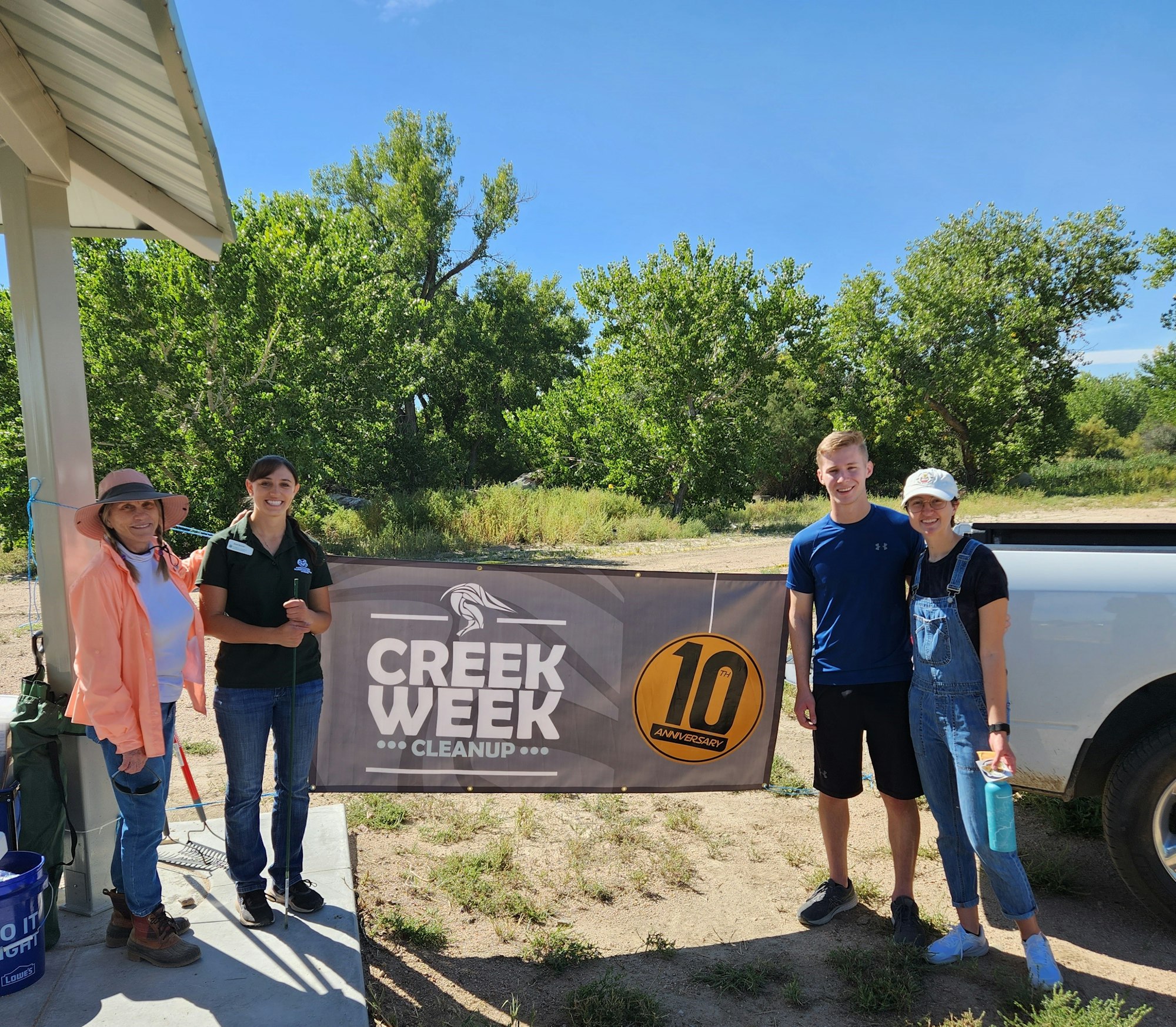 Four volunteers posing with a "Creek Week Cleanup" banner and cleanup tools in Confluence Park, Pueblo, CO.