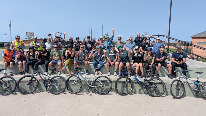A group of people with bicycles, wearing helmets, sitting on concrete steps and smiling outdoors.