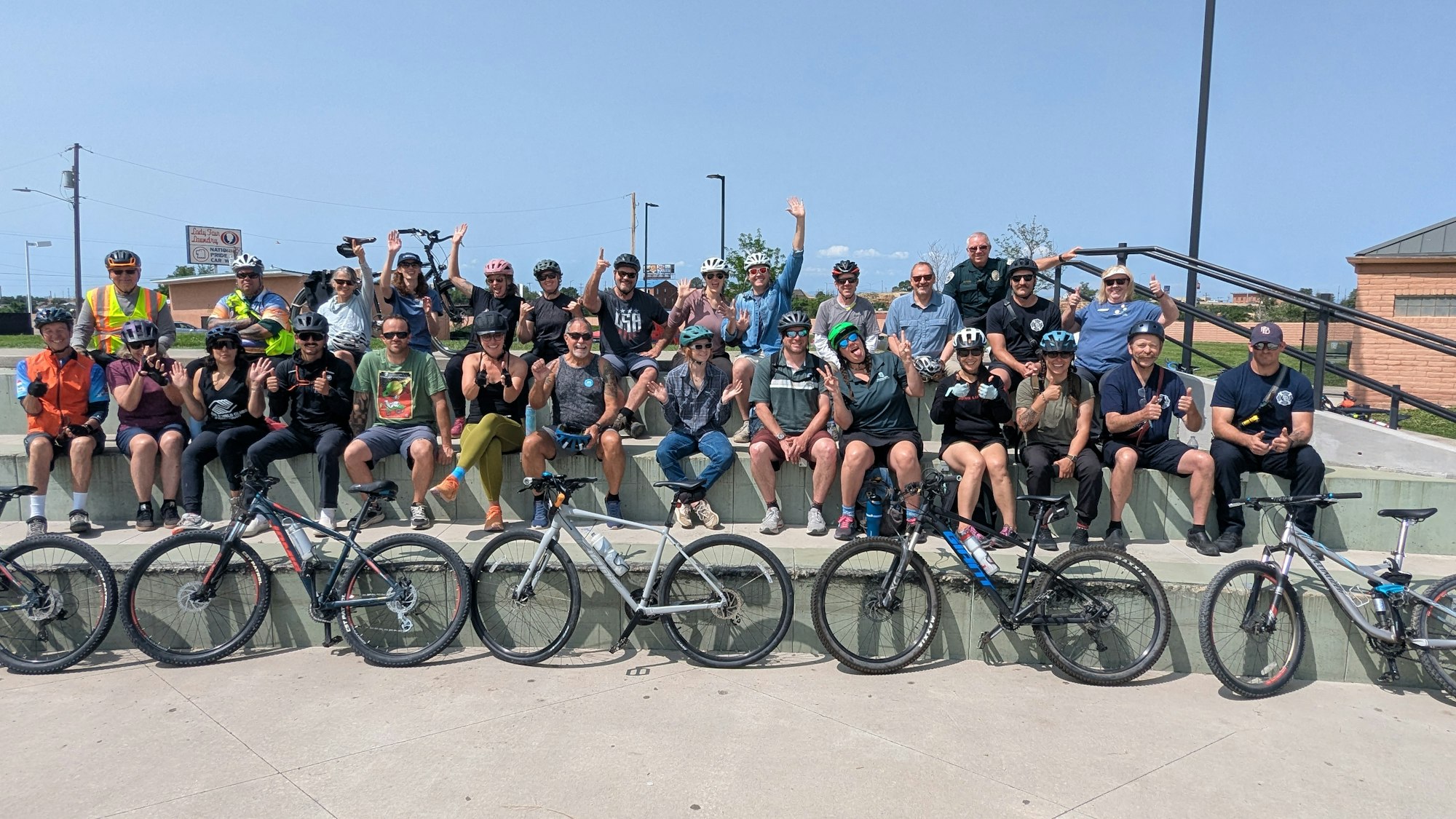 A group of people with bicycles, wearing helmets, sitting on concrete steps and smiling outdoors.