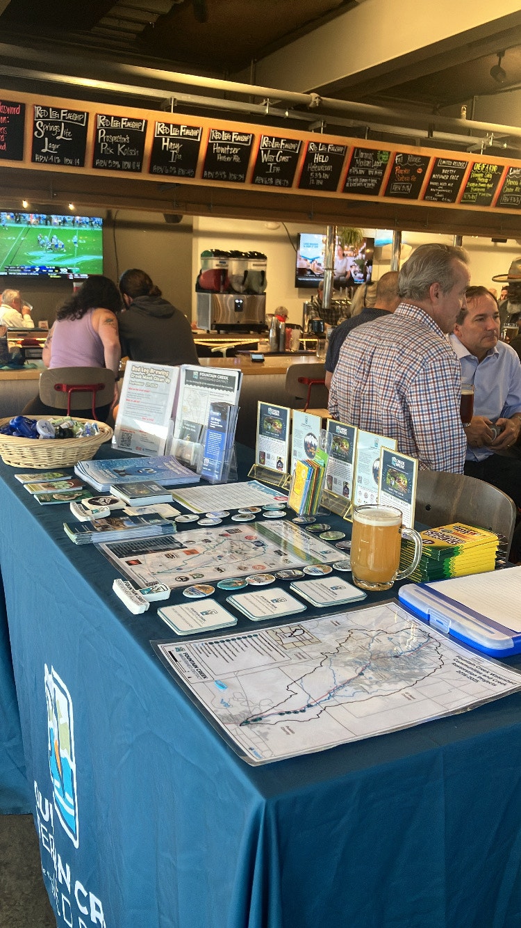 A bar scene featuring a promotional table with maps, brochures, and a beer, alongside patrons enjoying their drinks.