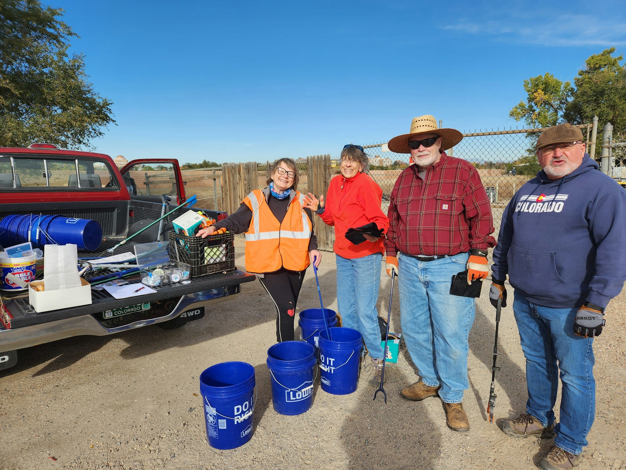 Four people in outdoor gear with cleaning tools and blue buckets beside a pickup truck.