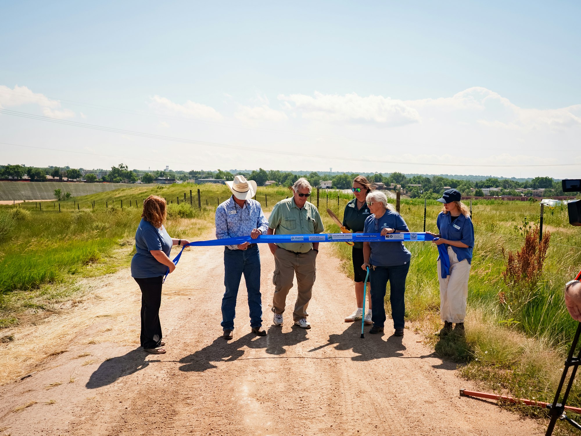 A group of people at a ribbon-cutting ceremony on a dirt path, celebrating completion of the Southmoor Dr restoration project in a grassy area under a clear sky.