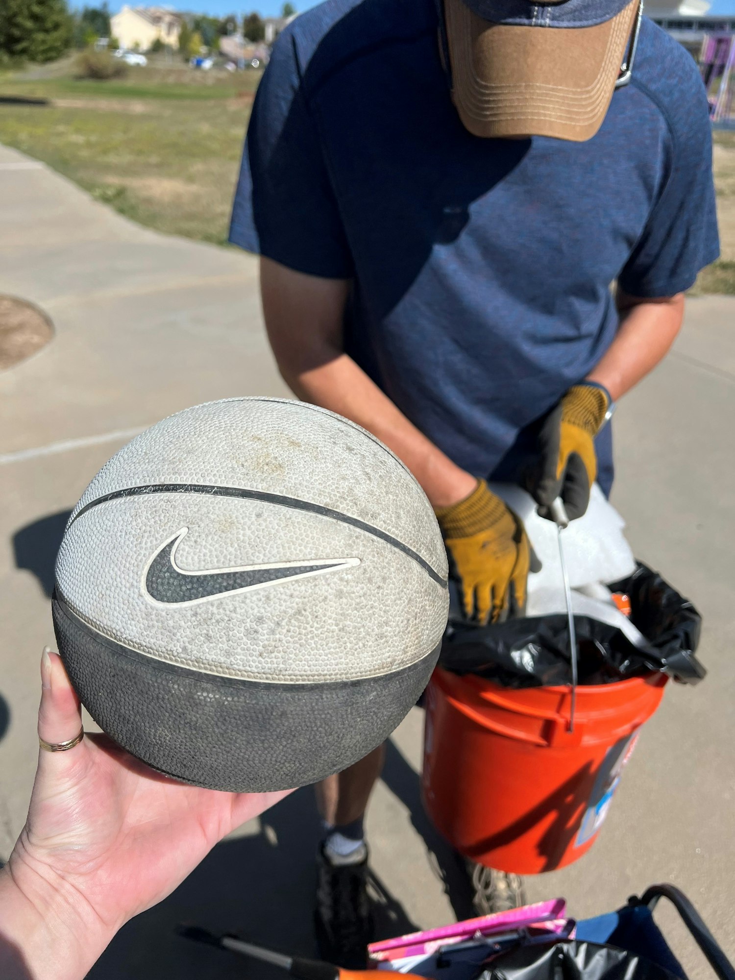 A person holding a basketball in the foreground; another person is picking up trash with a bucket and grabber tool.