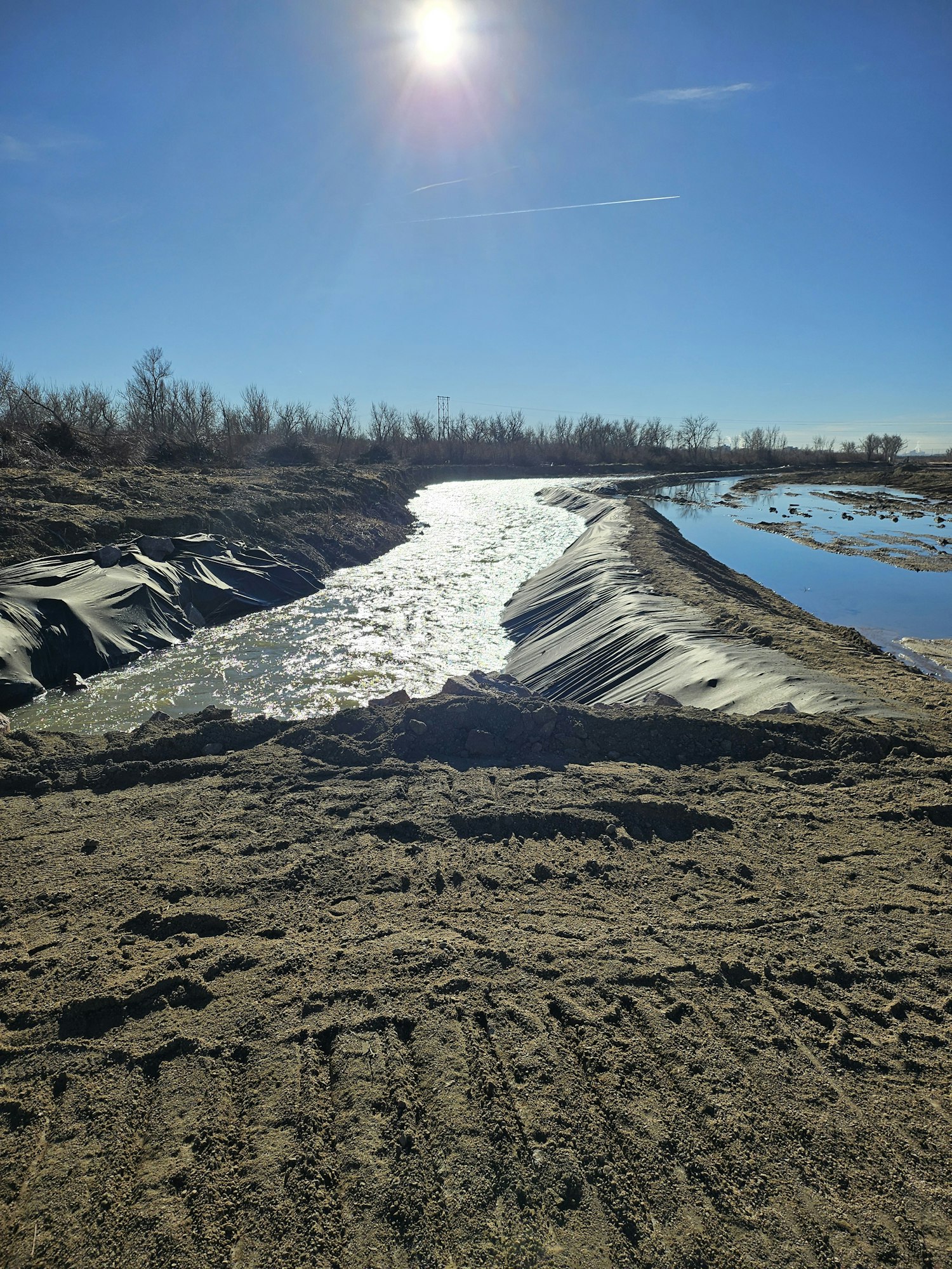 A river flows between two muddy banks with the sun shining in a clear blue sky.