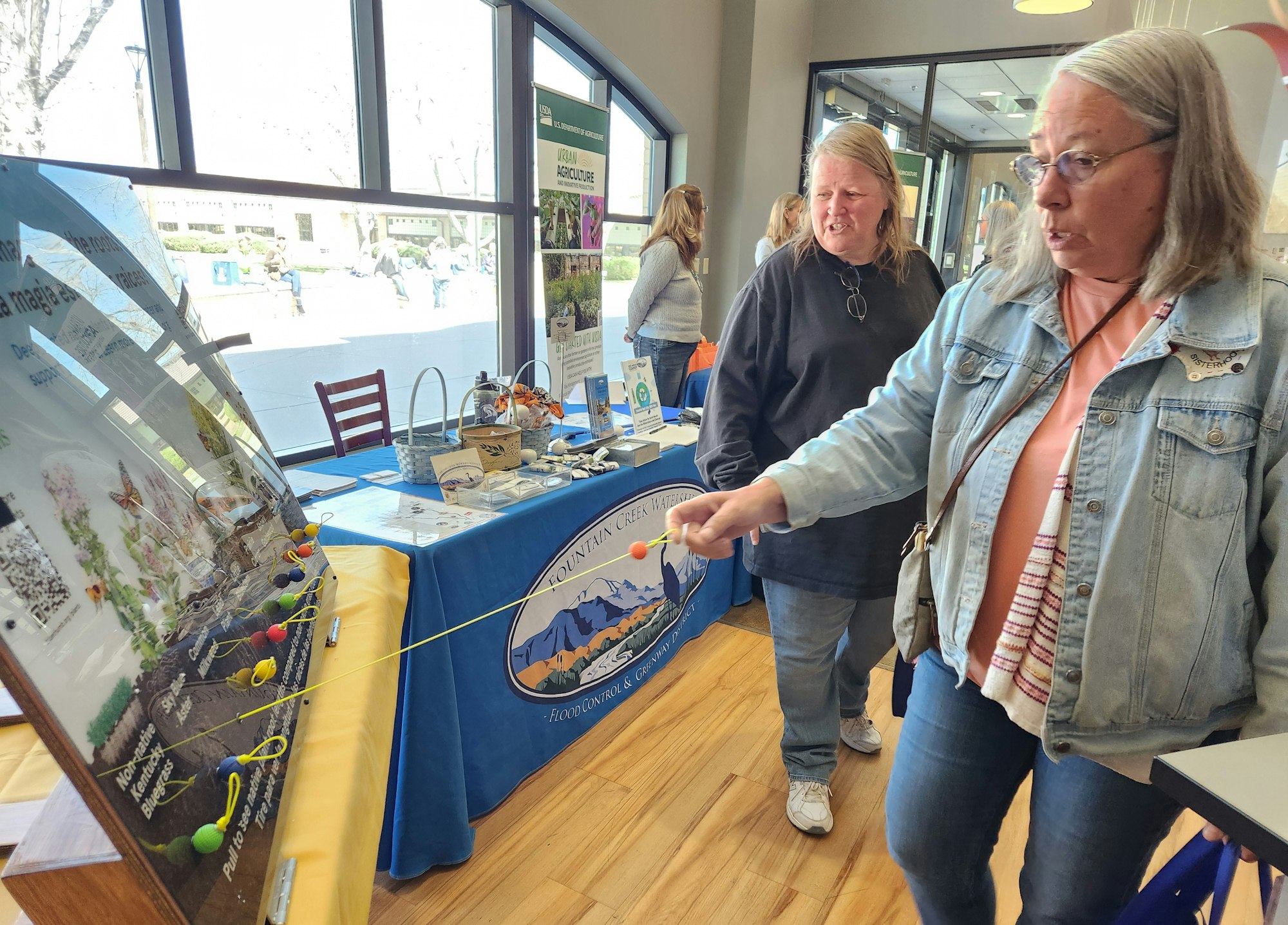 Two women interact with a display at an agricultural event. One is using a tool on a board, with informational booths in the background.