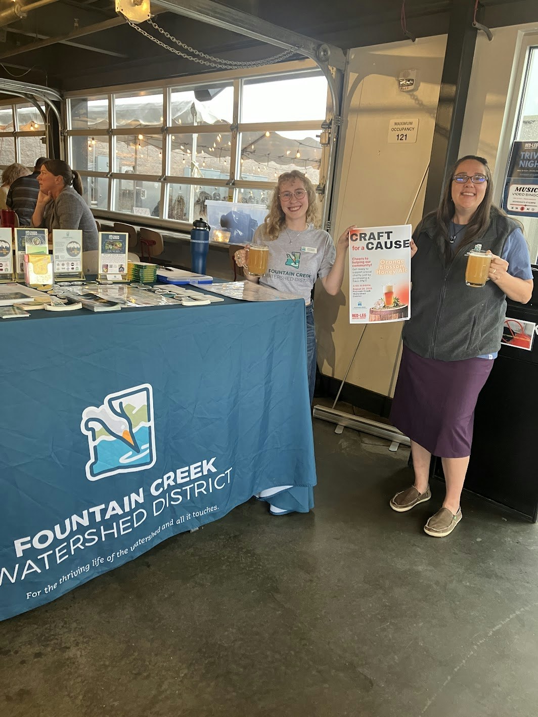 Two women hold drinks and a poster at a booth for the Fountain Creek Watershed District, promoting a craft event.