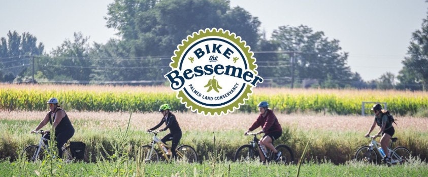 Cyclists riding through fields with "Bike the Bessemer" logo for Palmer Land Conservancy.