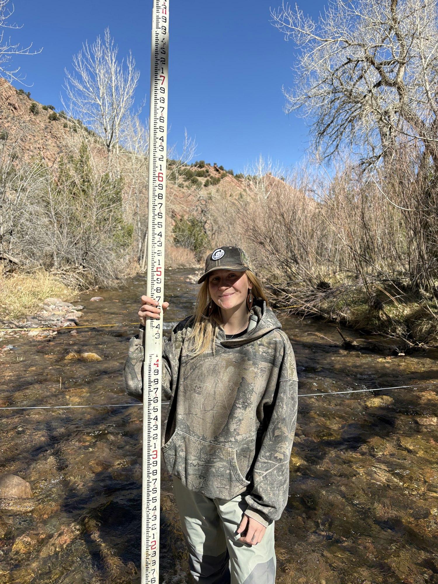 A person in camouflage stands by a stream, holding a measuring stick with markings, under a clear blue sky.