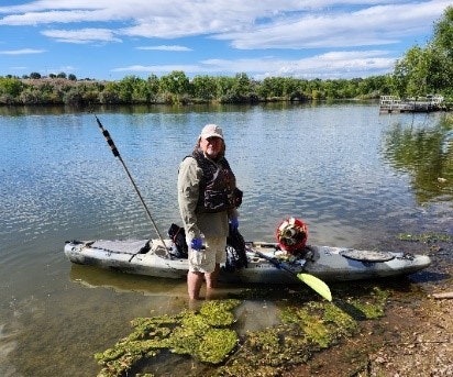 A person stands in a kayak near a riverbank, dressed for kayaking, with a paddle in hand and a dog nearby.