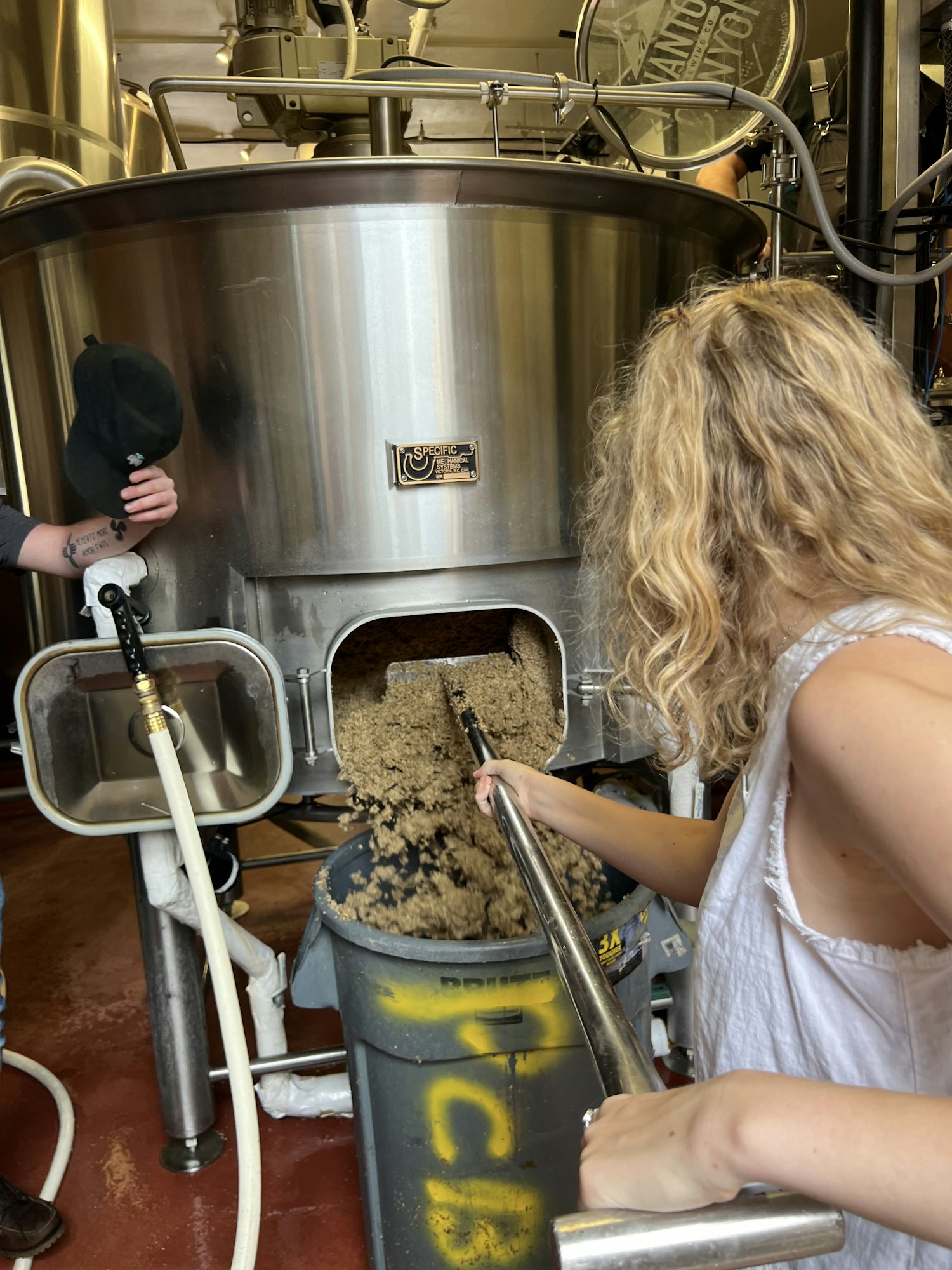 A person is using a large tool to transfer grain from a brewing vessel into a container in a brewery setting.