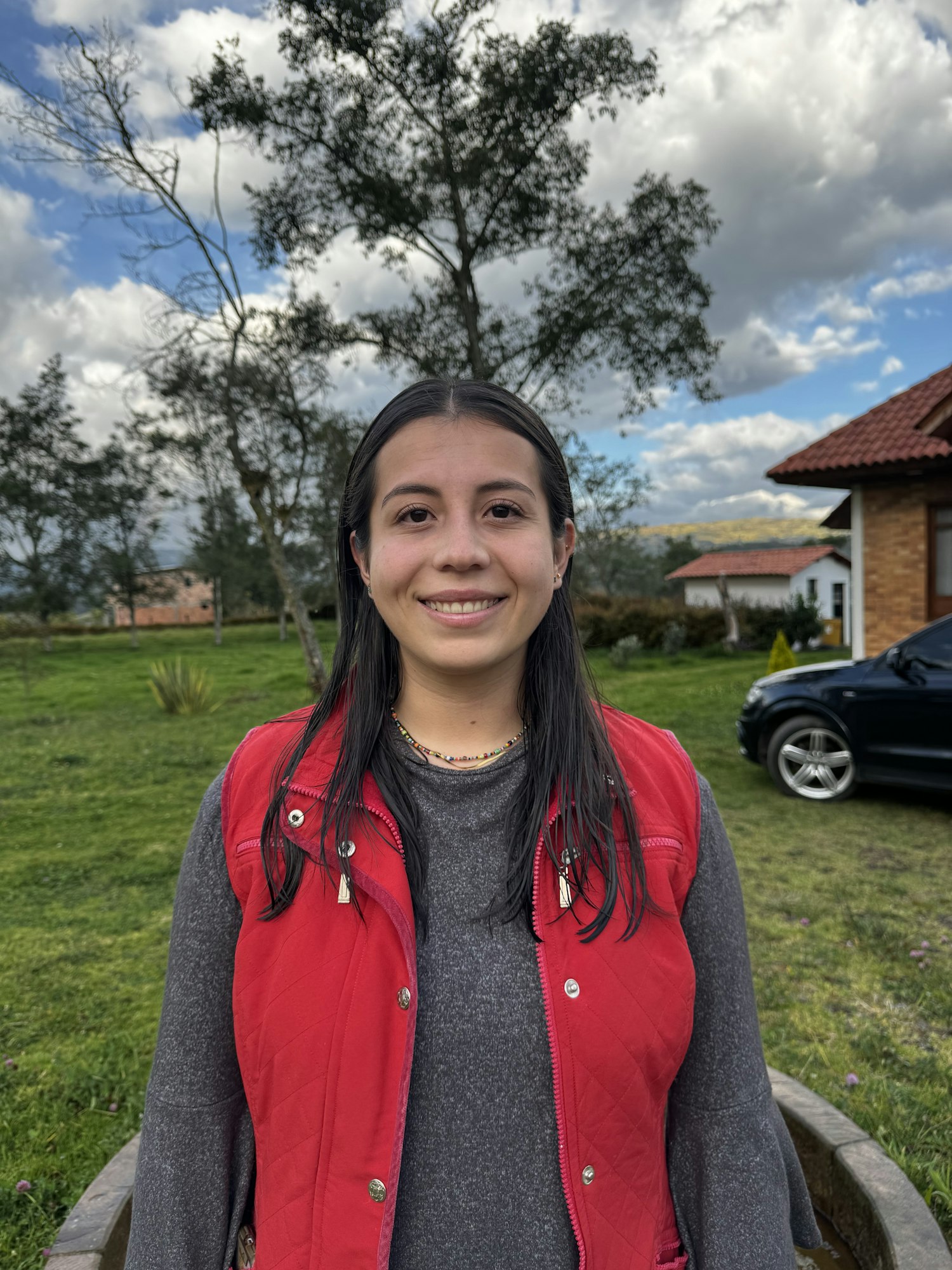Image of young woman in grey long sleeve and red vest standing outside with trees, grass, and buildings in the background.