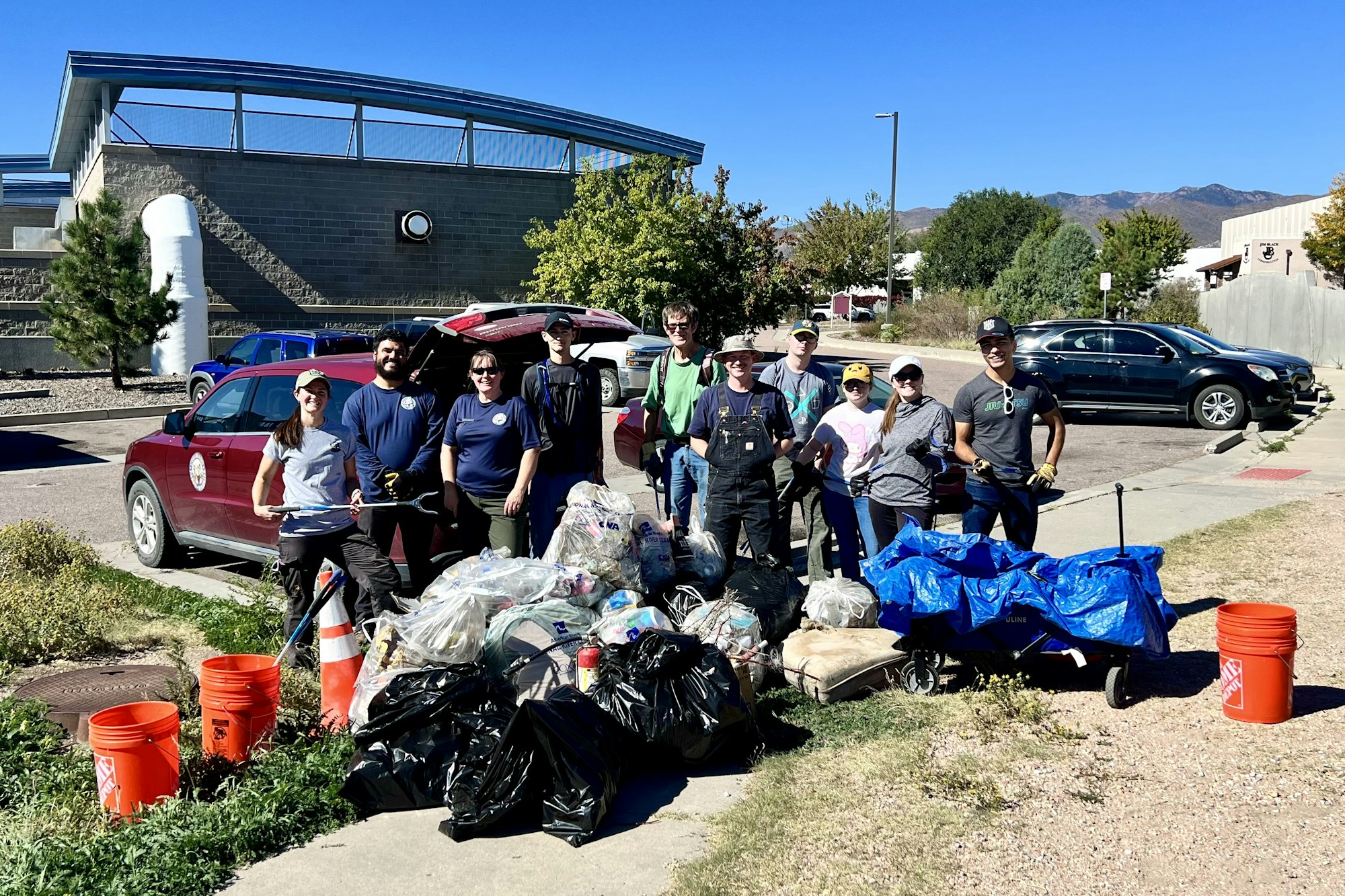 A group of people poses with collected trash bags and cleanup tools in a parking lot.