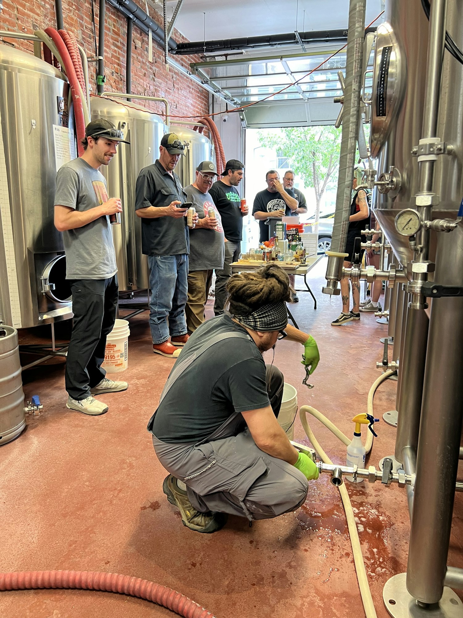 A group of people in a brewery, some watching while one person works on equipment, likely involved in beer brewing.