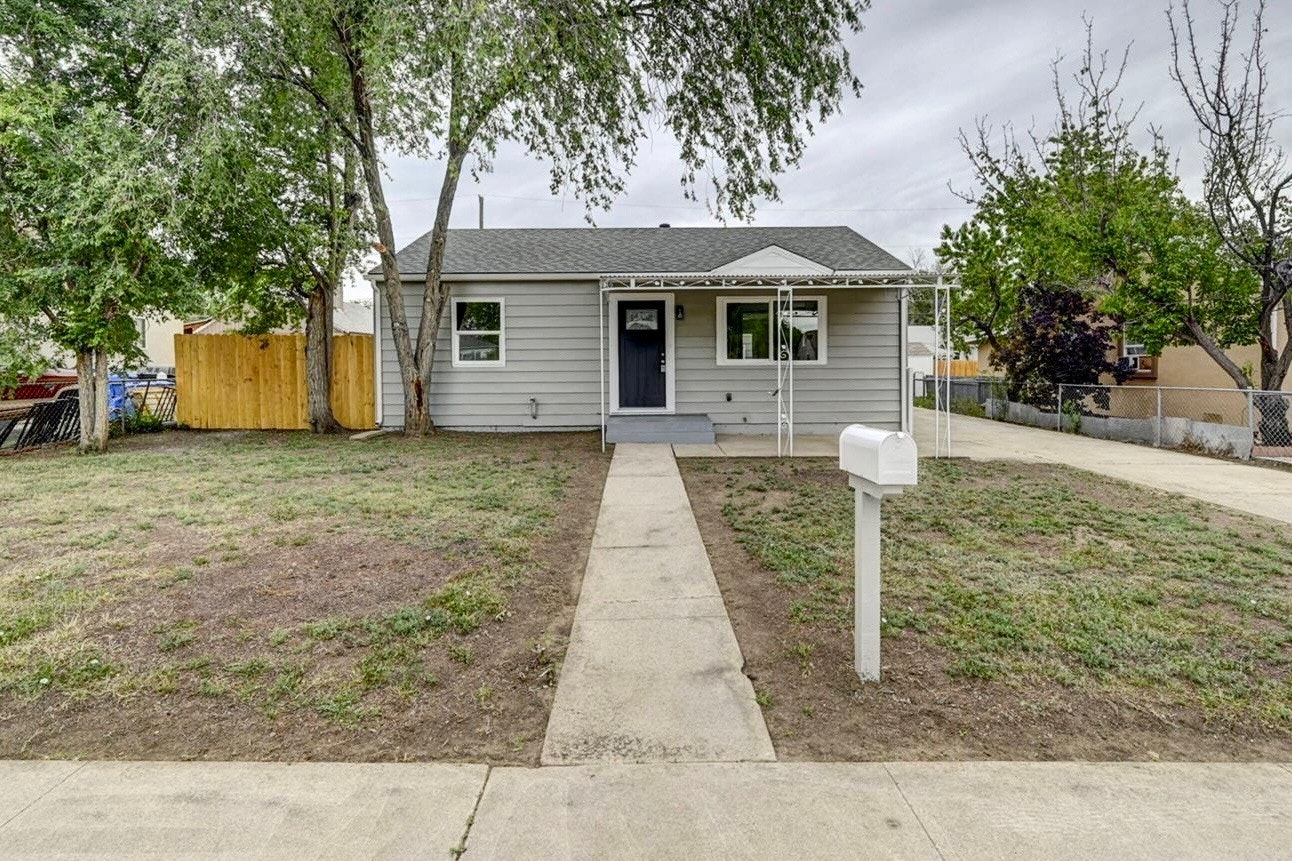 Before installation of a residential raingarden; photo of single-story gray house, small front yard, concrete path, white mailbox, trees on both sides, wooden fence in background.