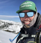A person in winter gear smiles for a selfie in a snowy mountain landscape, wearing sunglasses and a green cap.