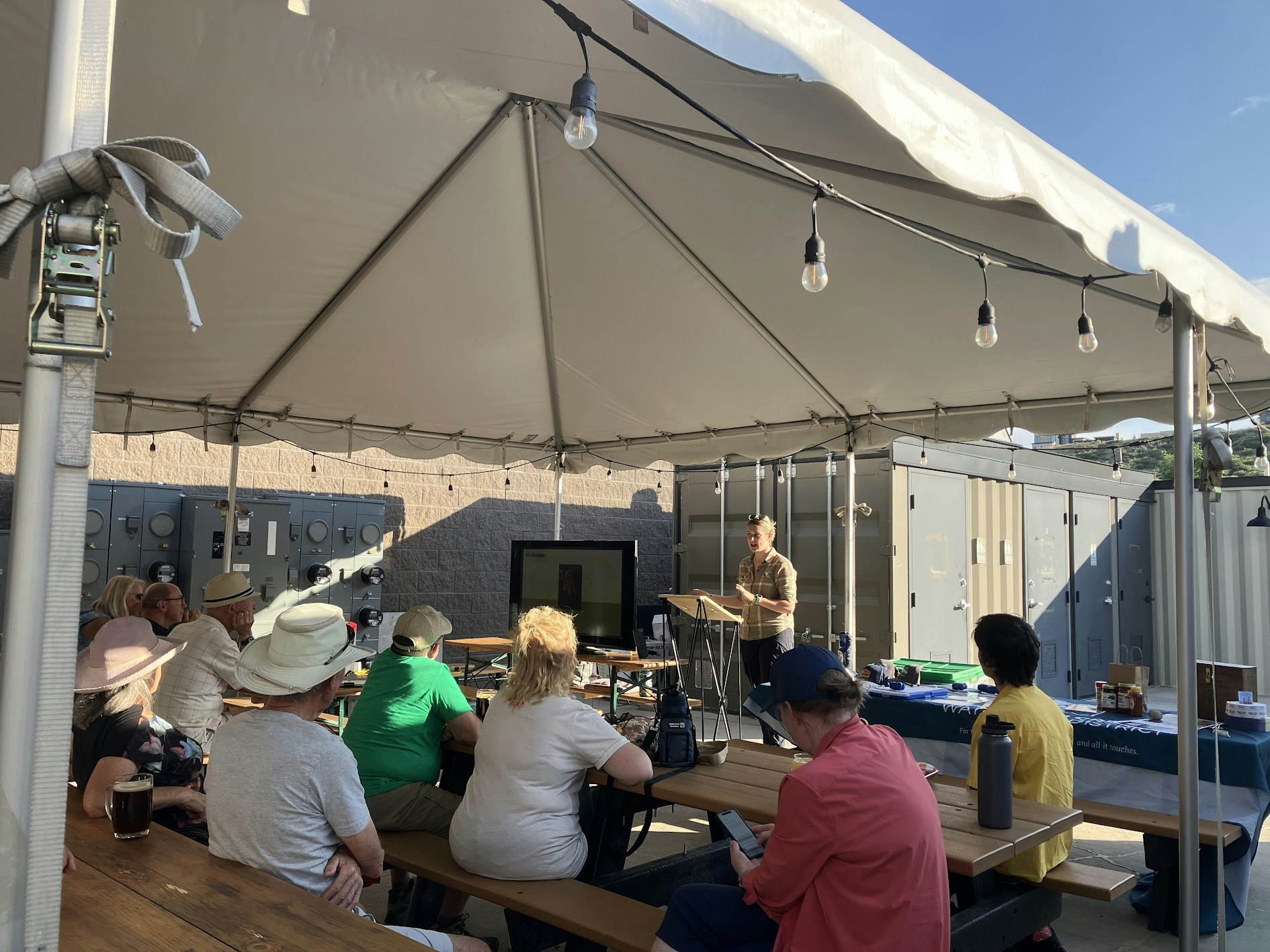 A group of people listens to a speaker under a tent, with tables and outdoor storage units in the background.