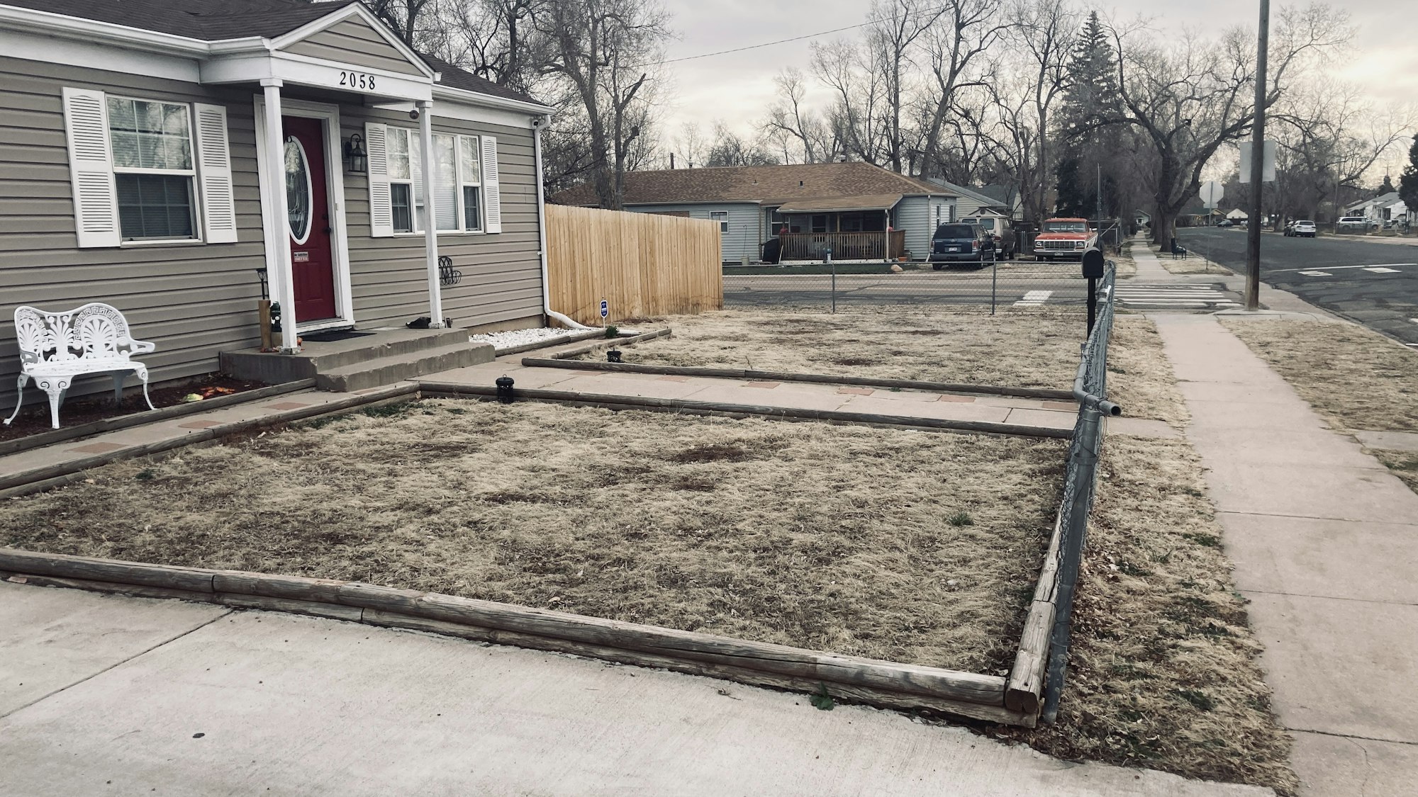 A single-story house with beige siding, white trim, a red door, and an empty front yard with a timber border. seen before installing a raingarden in a Stratton Meadows neighborhood