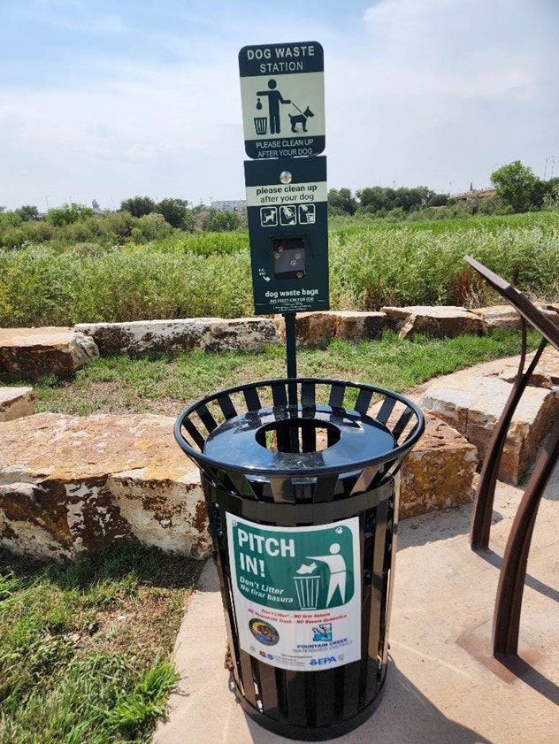 A dog waste station with a sign and dispensers for bags, next to a trash can labeled "PITCH IN!" outdoors.