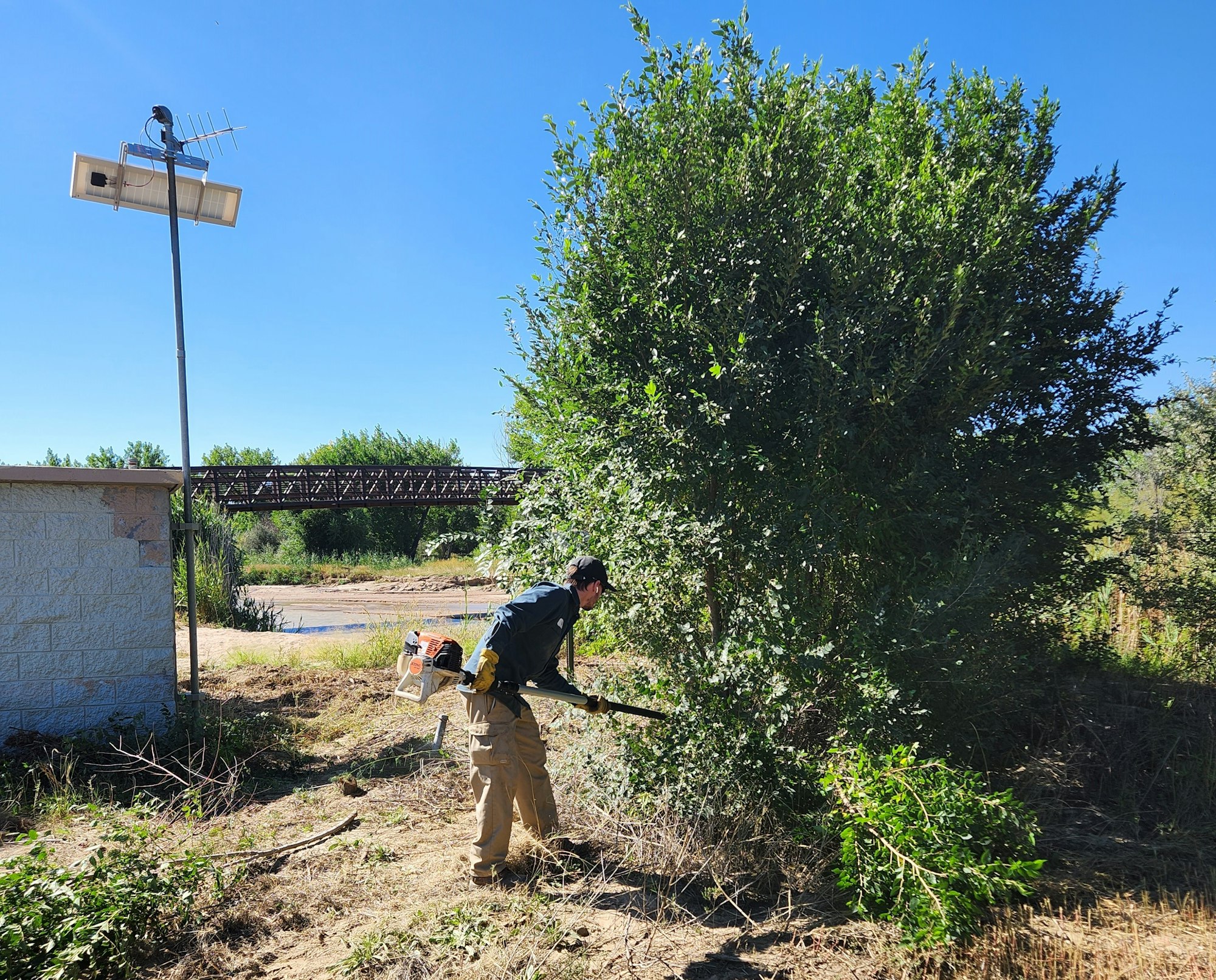 Person trimming bushes and clearing brush from around a water monitoring station with a solar panel near a bridge in Pueblo, CO.
