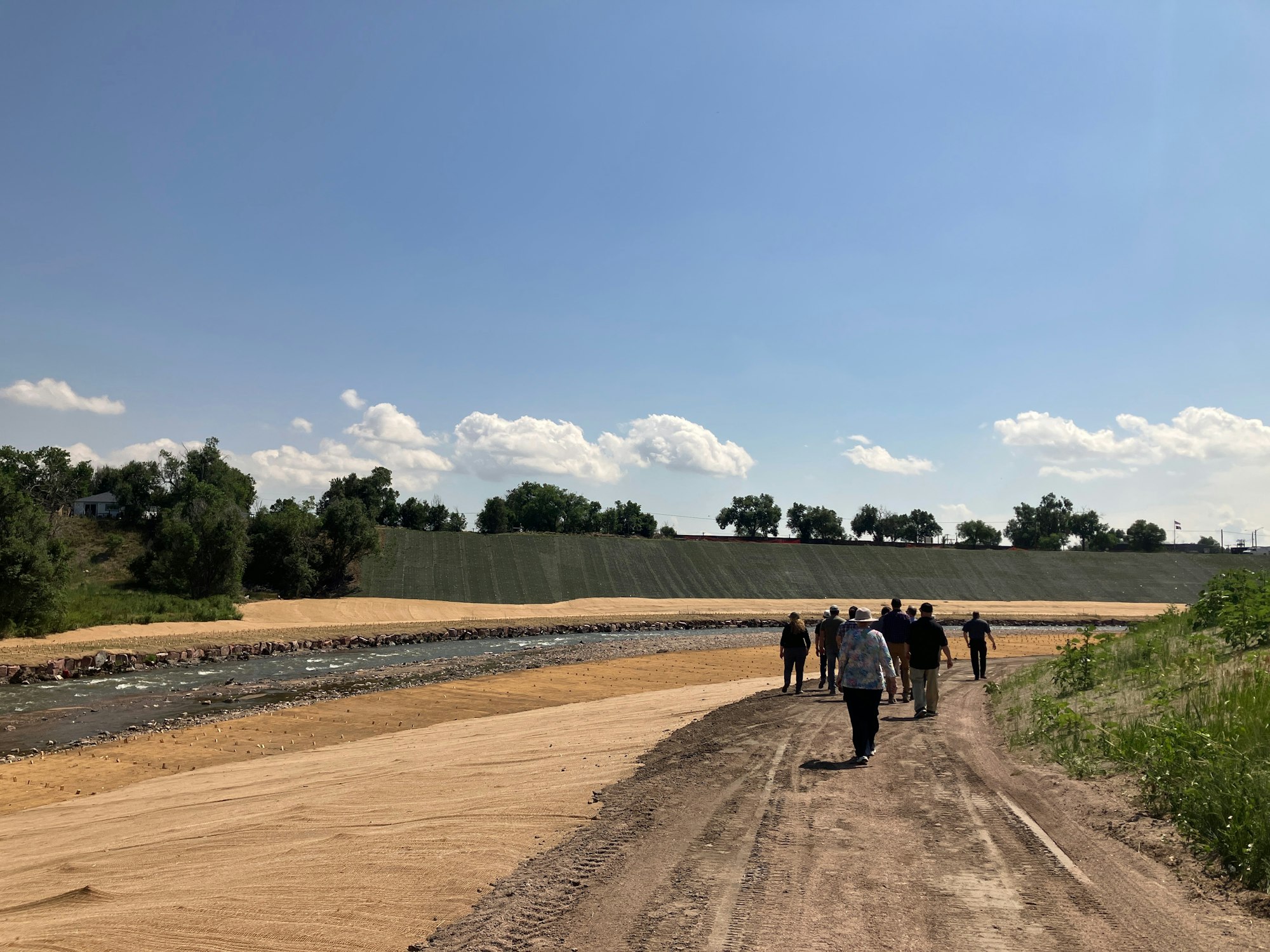 A group of people walking along a dirt path beside a river with trees and a grassy slope under a clear blue sky.