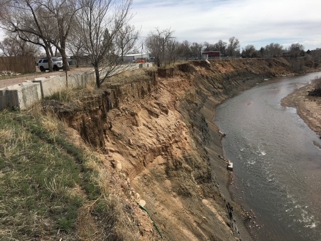 Eroded riverbank near a road with vehicles and trailers, indicating possible environmental issues.