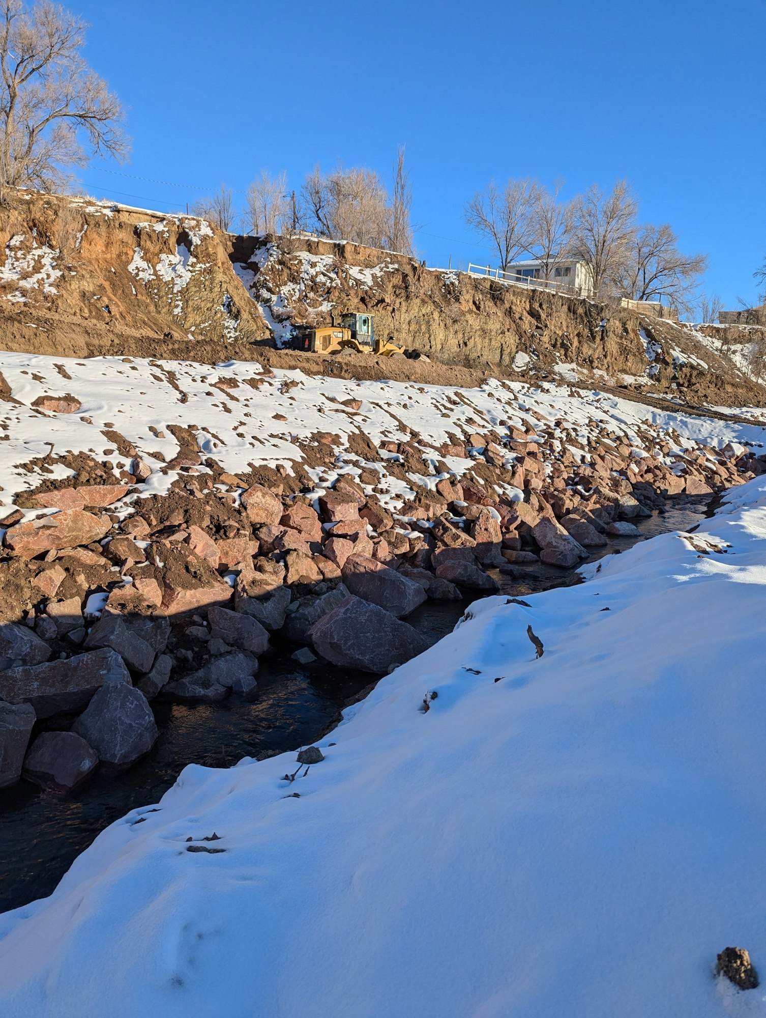 A snowy slope with a river, rocks, and a construction vehicle; trees and a house on a cliff above.