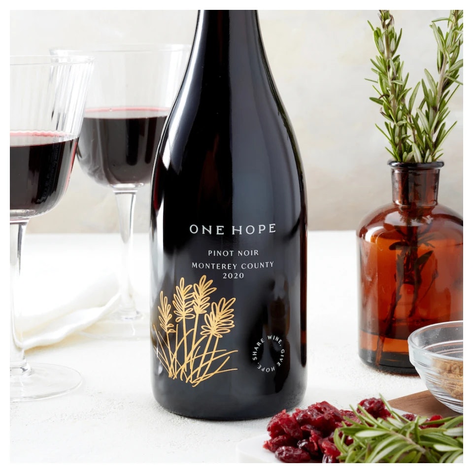 A bottle of wine labeled "ONE HOPE Pinot Noir 2020," two glasses, a brown bottle with greenery, and food on a table.