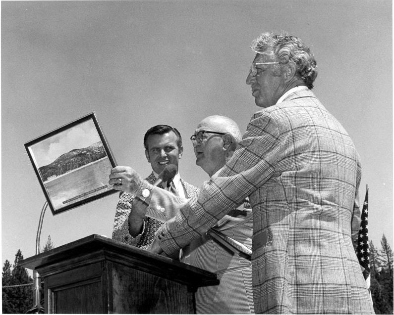 Three men in suits at a podium, one holding a framed picture, with an American flag visible in the background.