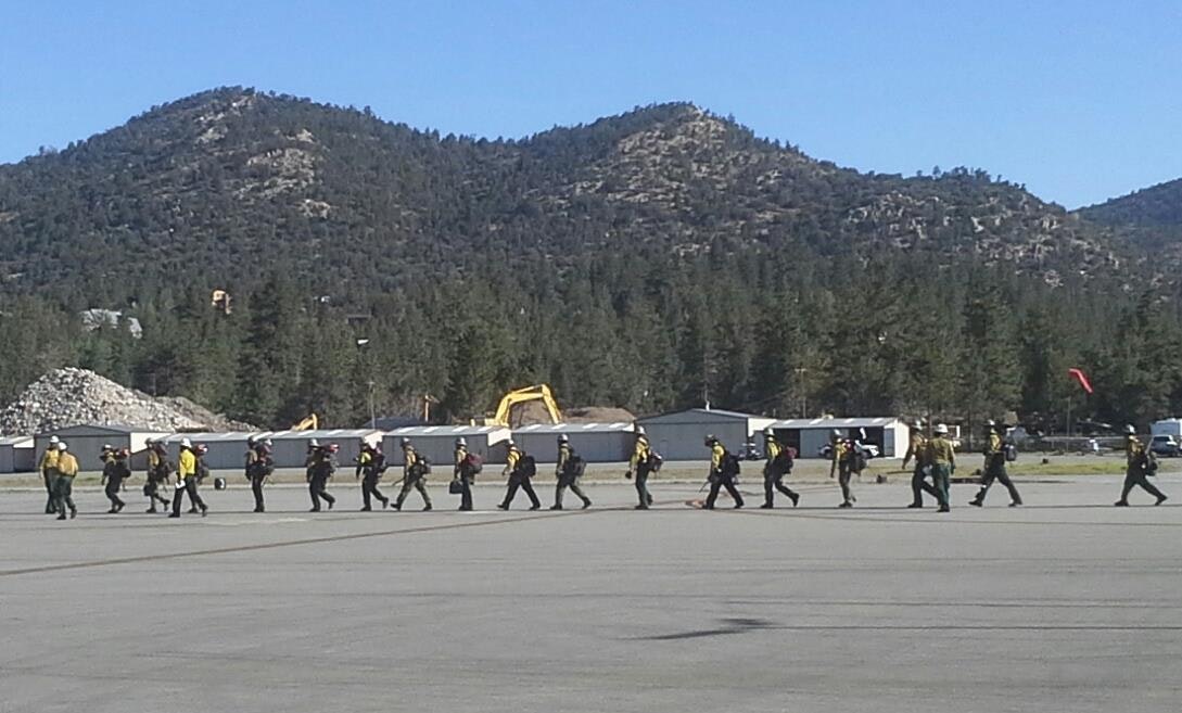 A line of people in uniforms walking on a tarmac with mountains and trees in the background.