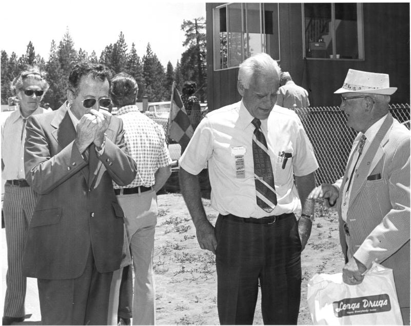 A group of men in suits outdoors, one playing harmonica, another holding a "Longs Drugs" bag, near a building and fence.