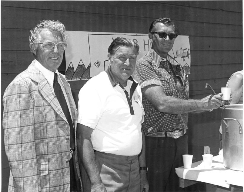 Three men standing outdoors, one pouring a drink from a large container. They are in front of a sign and near a table with cups.