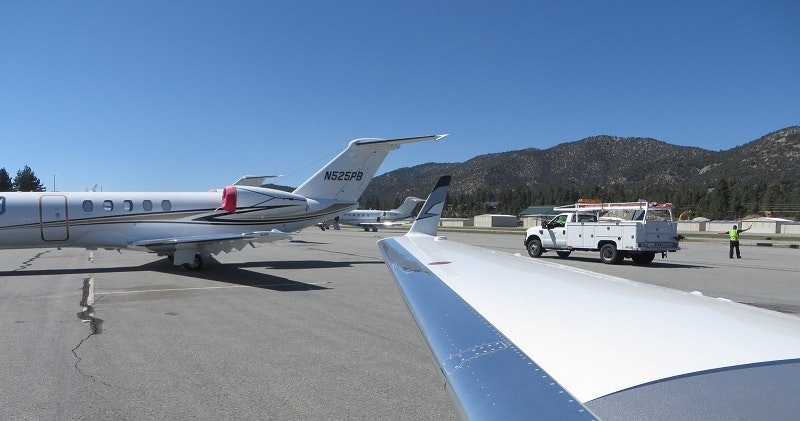 Parked private jets, airport ground crew, truck, and mountains in the background under clear blue sky.
