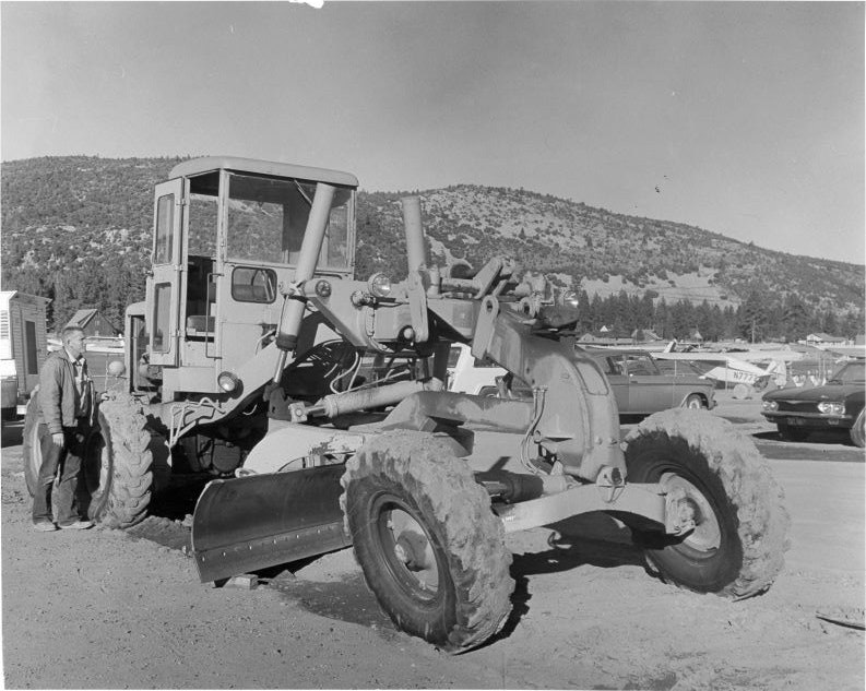 A person stands next to a large construction vehicle, possibly a road grader, with hills and cars in the background.