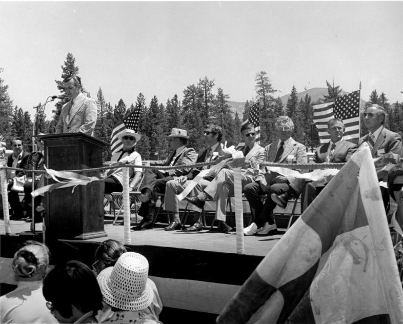 A man speaks at a podium outdoors; a seated audience with American flags is in the background.