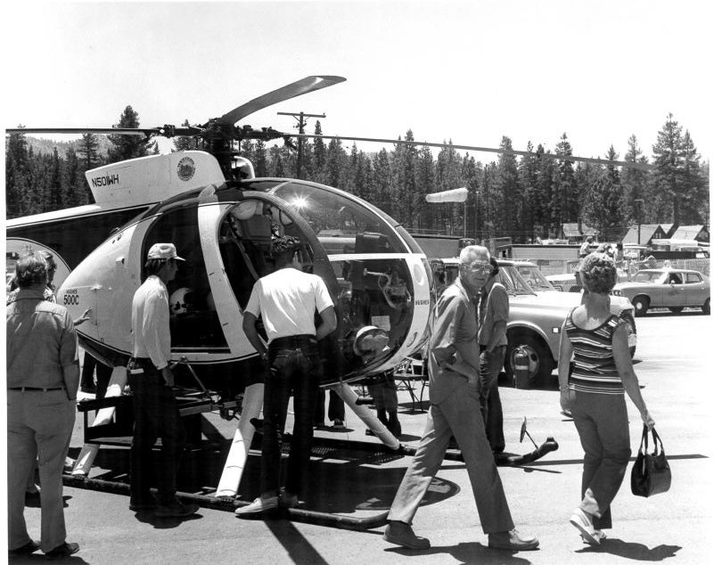 A group of people observing a helicopter on the ground, with trees and parked cars in the background.