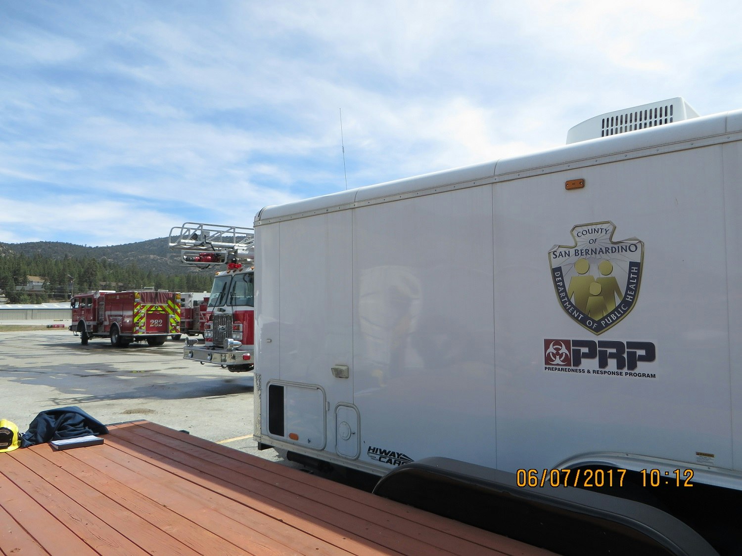 Fire trucks and a public health trailer in a parking area under a cloudy sky.