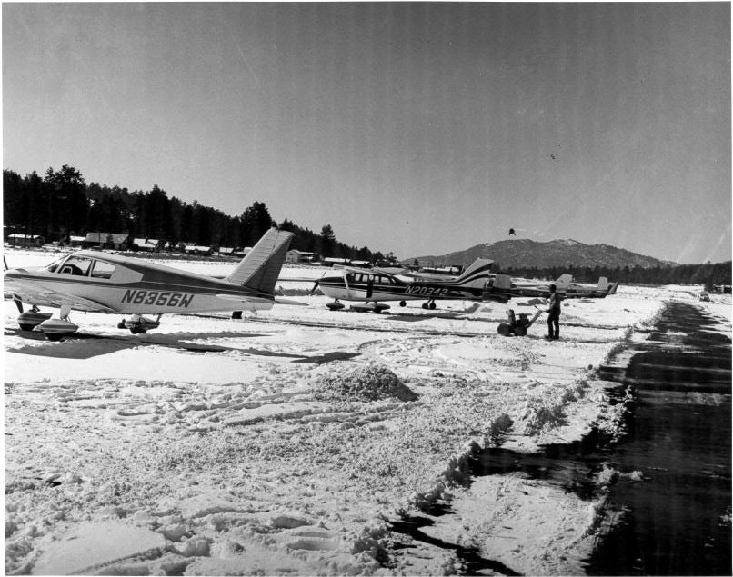 Small aircraft parked on a snow-covered runway, with a person nearby and trees in the background.