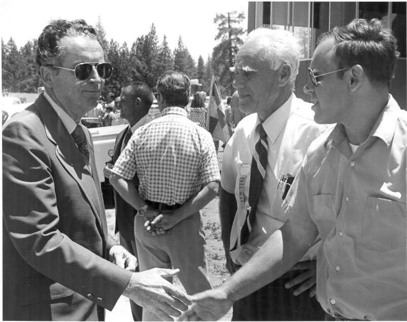 Three men are shaking hands and talking outdoors, with others standing in the background.