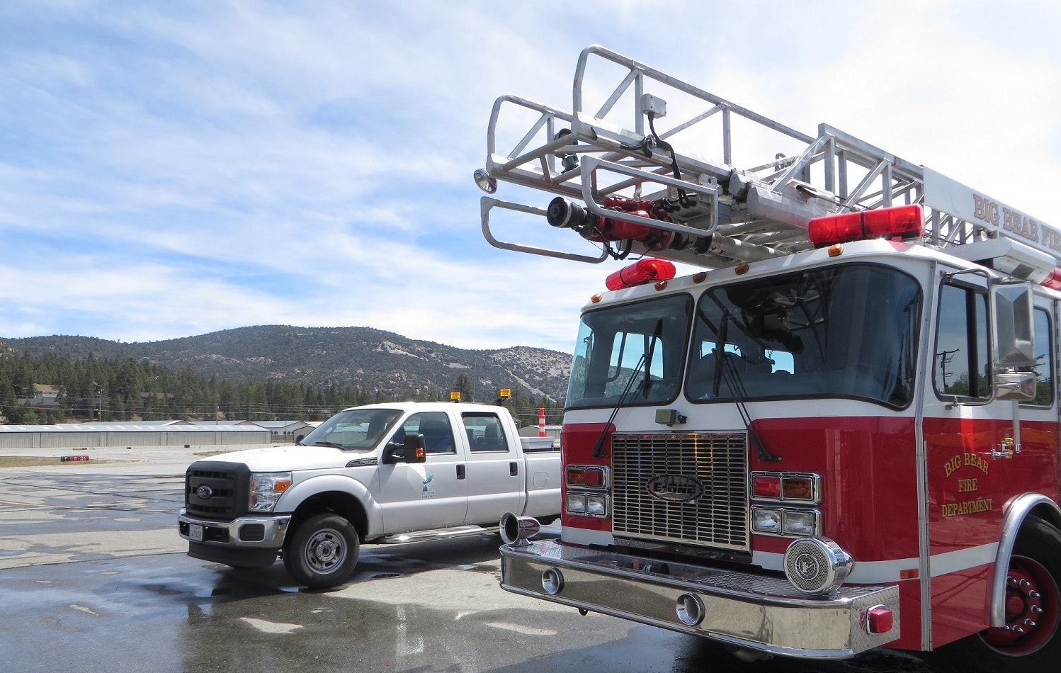 A fire truck and a white pickup truck parked on a tarmac with mountains and trees in the background.