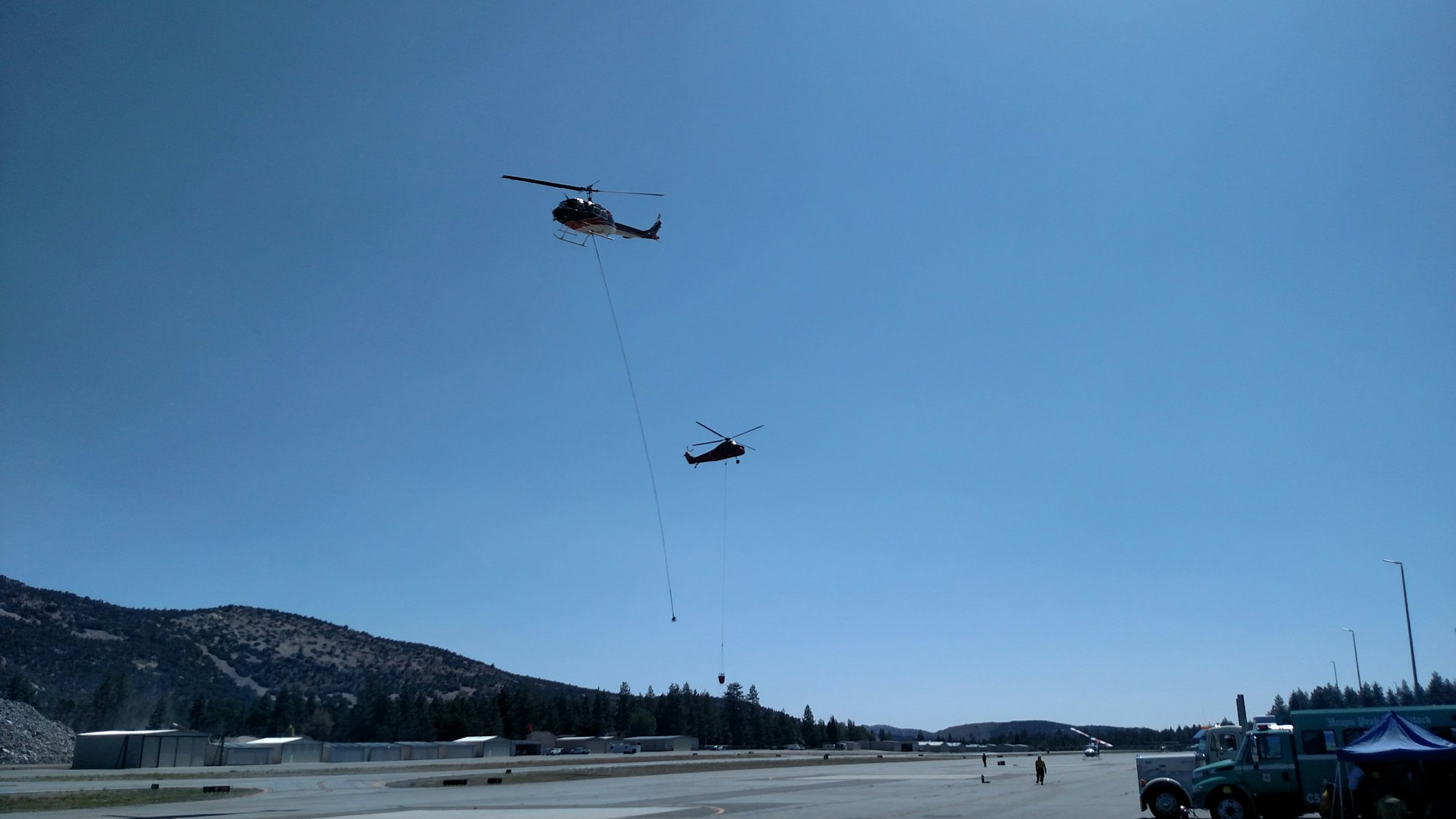 Two helicopters are flying over an airfield, carrying equipment on long lines.