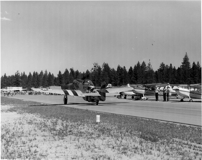 A vintage plane taxis on a runway with several small aircraft parked nearby, trees in the background, and a small crowd on the side.