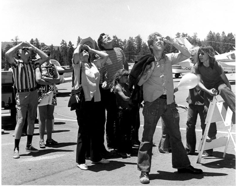 A group of people is looking up at the sky near an airplane. One person holds a balloon.