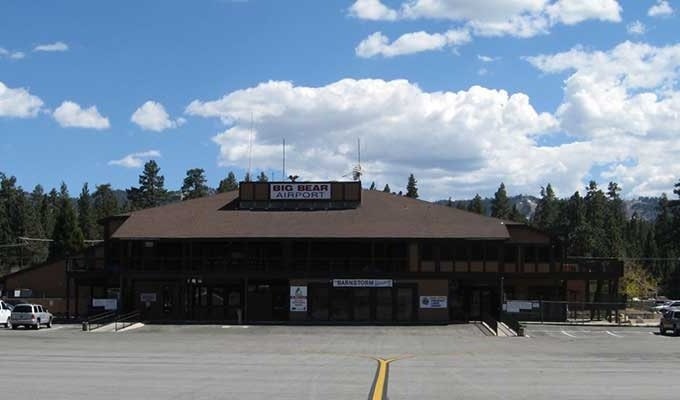 A small airport building labeled "Big Bear Airport" with a parking area, surrounded by trees and a cloudy sky.