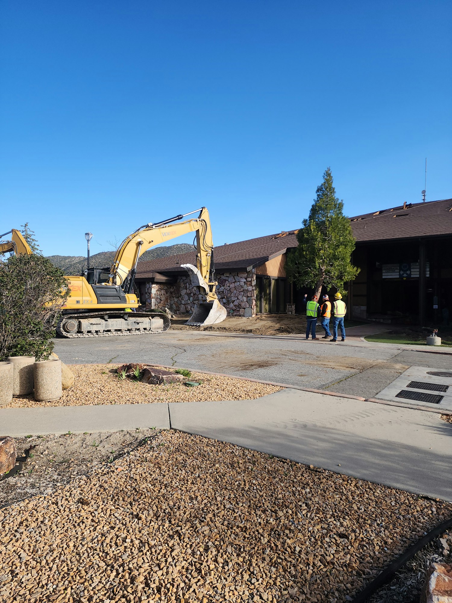 Construction site with an excavator, two workers in high-visibility vests, and a building in the background.