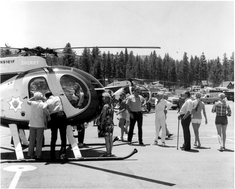 A group of people gathers around a sheriff helicopter in a parking lot, with more vehicles and trees in the background.