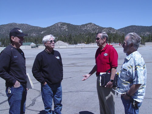Four men standing and talking outdoors on a paved area with mountains in the background.