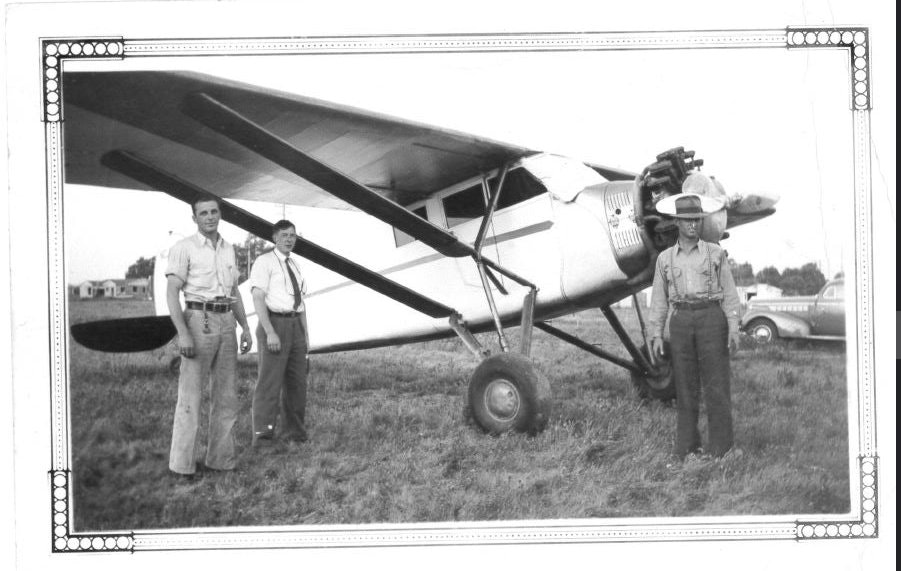 Three men stand beside a vintage propeller airplane on a grassy field, with cars in the background.