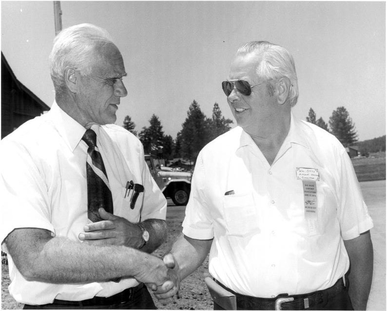 Two men shaking hands outdoors, both in white shirts, ties, and badges, with a background of trees.
