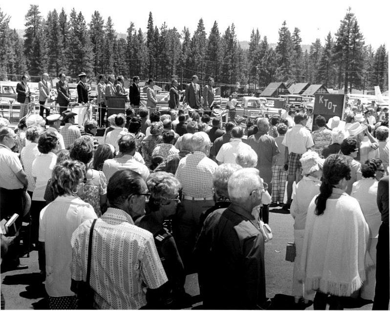 A crowd gathered outdoors near parked cars and a stage with people on it, surrounded by trees. A sign with "KTOT" is visible.