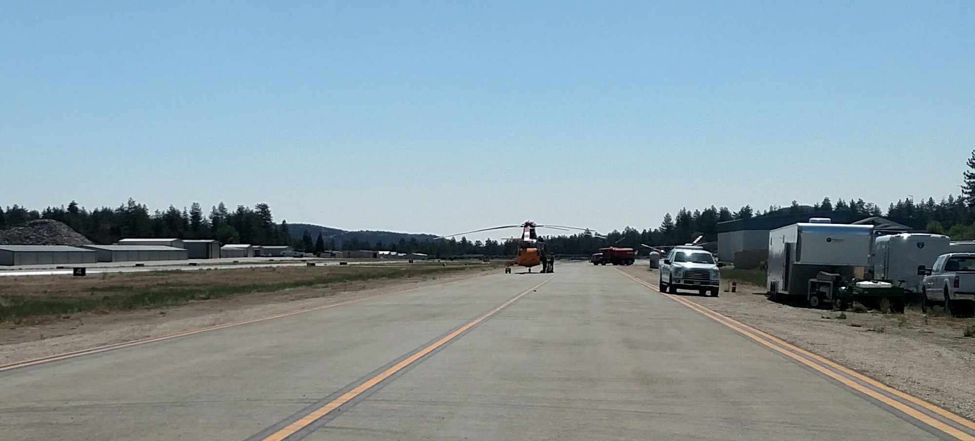 Helicopter on a runway, surrounded by vehicles and buildings, set against trees and a clear sky.